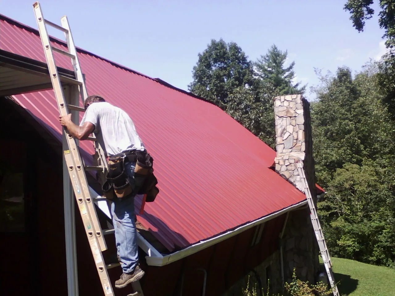 Roofer on a ladder beside a red metal roof and stone chimney — Soffit & Fascia Repair by Roofing Brothers in Kenvil